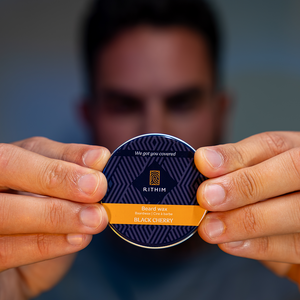 A person holds a round tin of Rithim Black Cherry Beard Wax in front of their face, with the product label clearly visible against a blurred background.