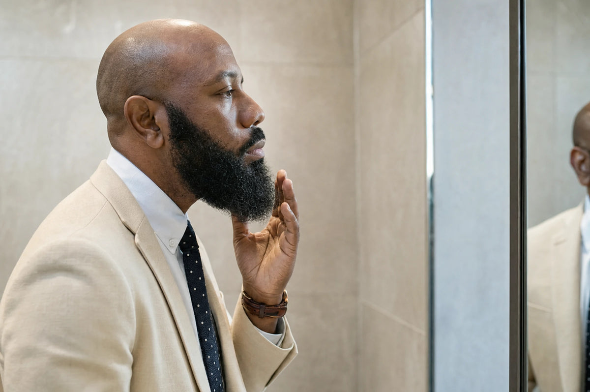 Bald man in a cream suit checking his full beard in a mirror