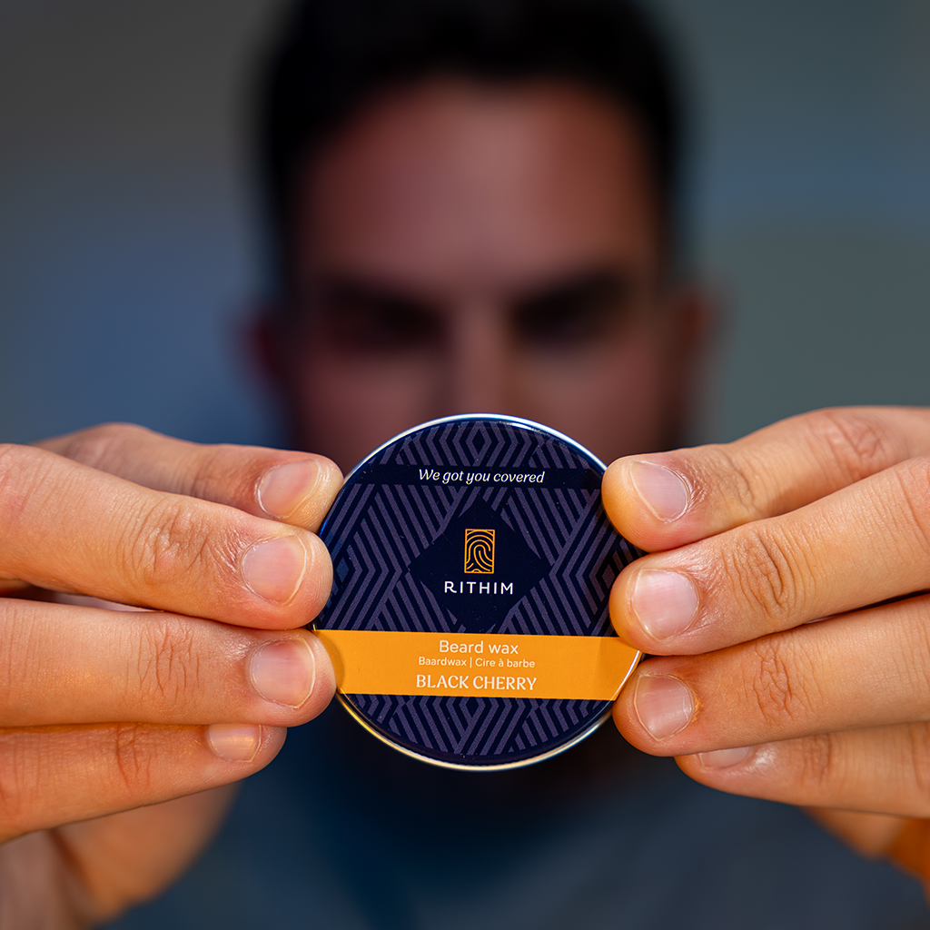A person holds a round tin of Rithim Black Cherry Beard Wax in front of their face, with the product label clearly visible against a blurred background.